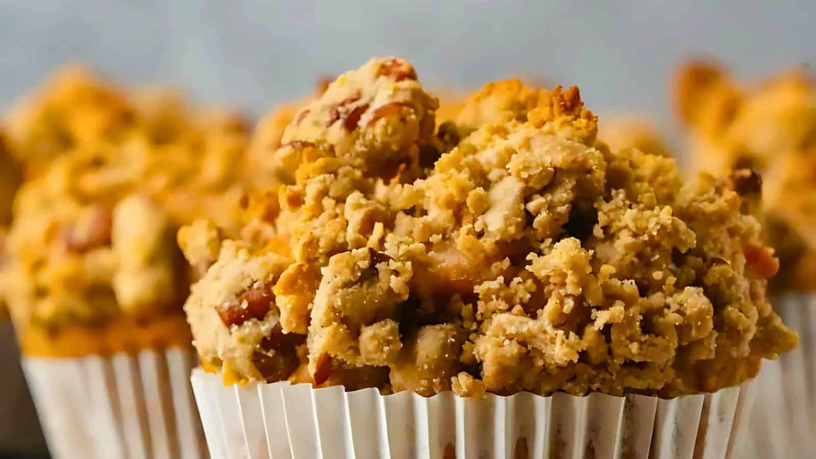 Close-up of a muffin with a crumbly topping in a white cupcake liner, with other muffins blurred in the background.