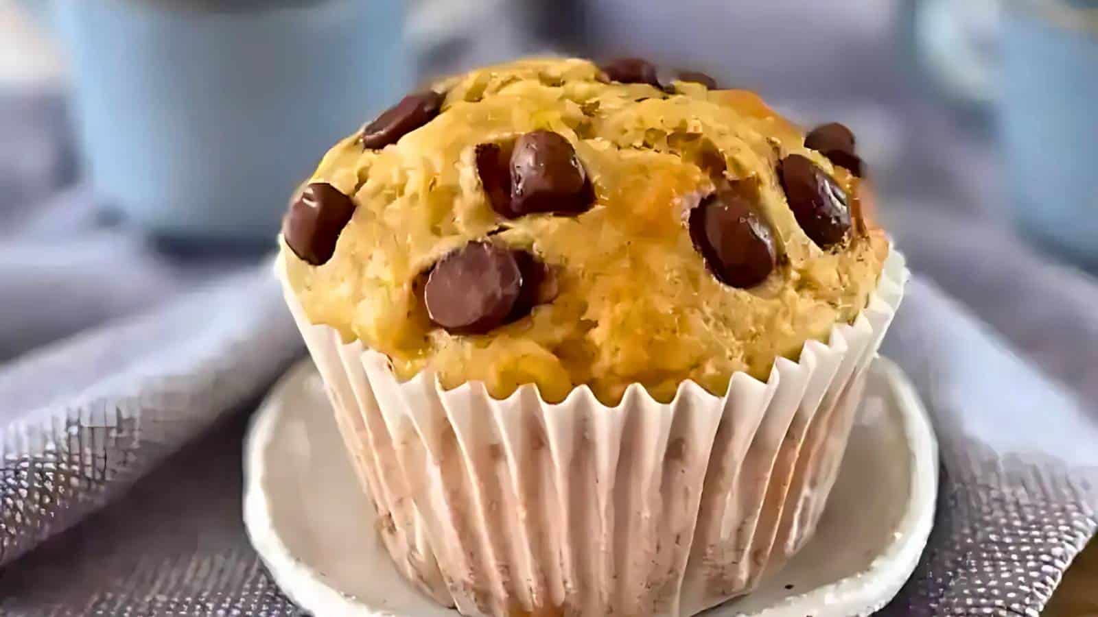 A chocolate chip muffin on a small white plate, surrounded by a textured cloth, with blurred blue cups in the background.