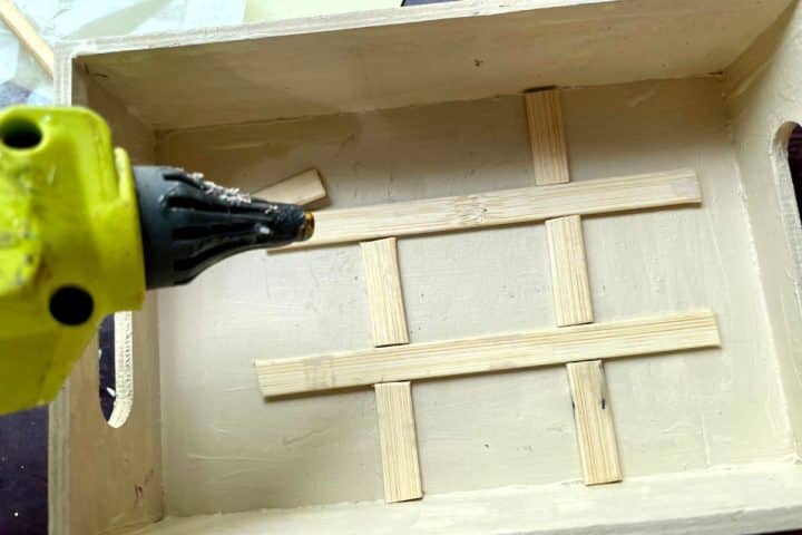 A hot glue gun is being used to glue intersecting wooden slats inside a white wooden box with handles.