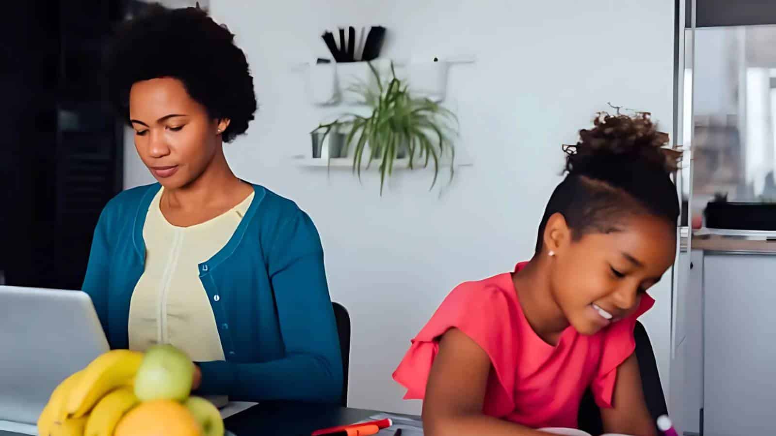 A woman works on a laptop while a girl writes at a table. A bowl of fruit and a plant are visible.