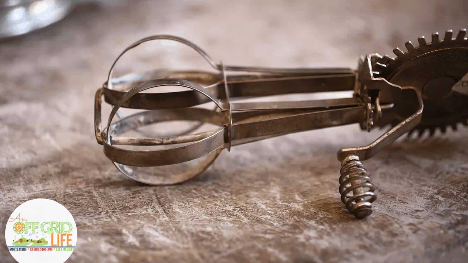 Close-up of a vintage egg beater on a textured surface, with a spiral handle and metal blades. Logo with text "Off Grid Life" in the bottom left corner.