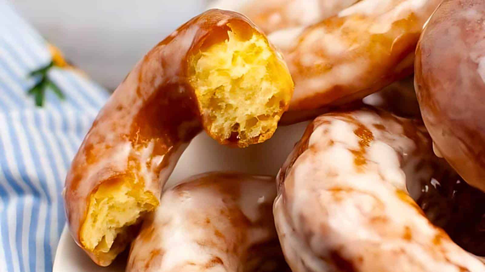 Close-up of glazed cruller donuts, with one showing a bite taken out, on a plate with a striped fabric in the background.