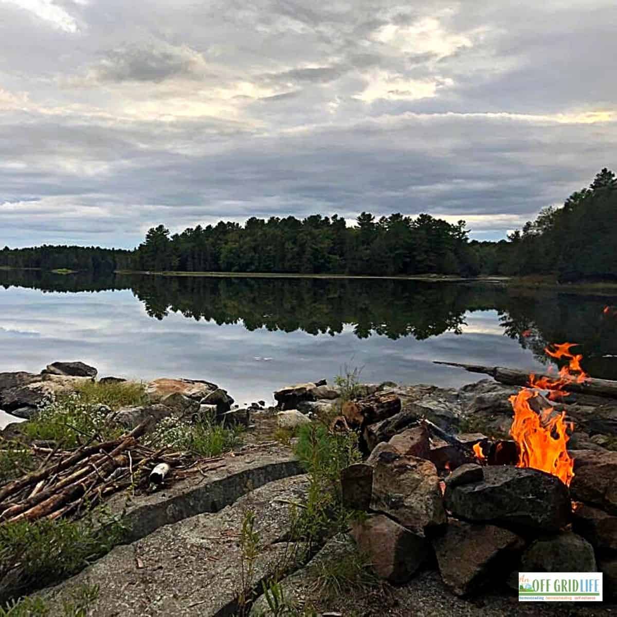 a campfire surrounded by stones next to a calm lake on a cloudy day.