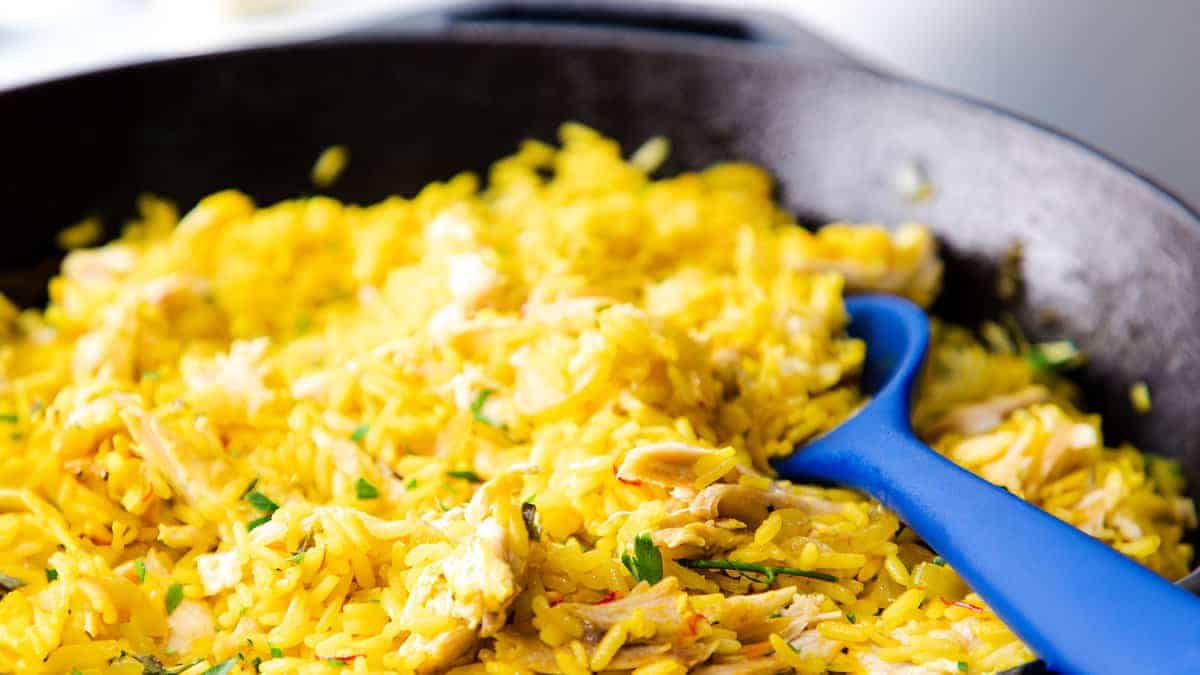 A skillet with yellow rice, shredded chicken, and herbs, served with a blue spoon.