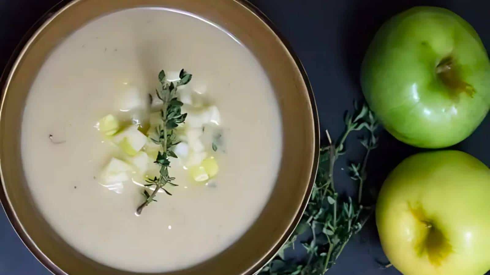 A bowl of creamy soup garnished with herbs and chopped ingredients, alongside two green apples and a sprig of thyme on a dark surface.