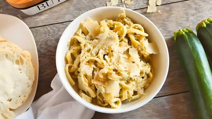 A bowl of pasta with grated cheese sits on a wooden table, paired perfectly with fresh zucchinis and a plate holding a slice of buttered bread—an ideal centerpiece for fall family feasts.