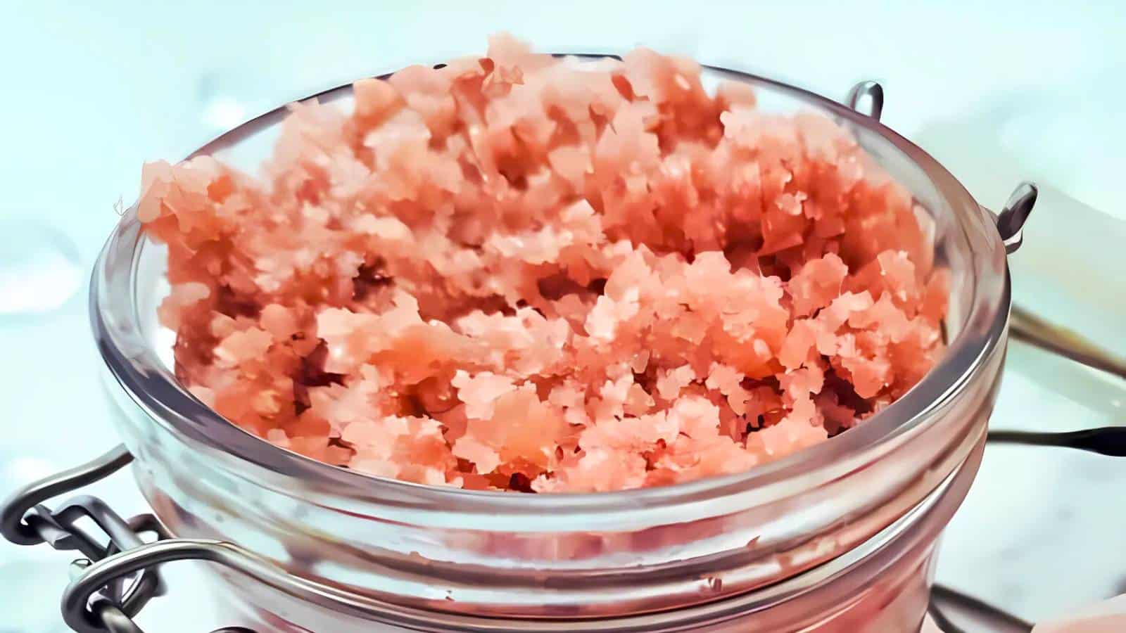 A clear jar filled with pink scrub, featuring a coarse texture, is shown with a metal latch closure on a softly blurred background.