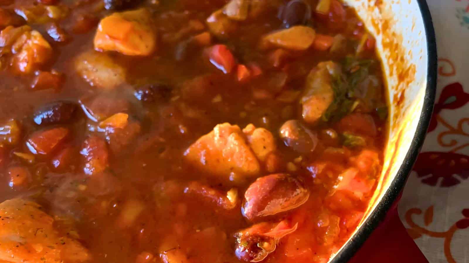 Close-up of a pot filled with thick, red stew containing chunks of chicken, tomatoes, beans, and herbs.