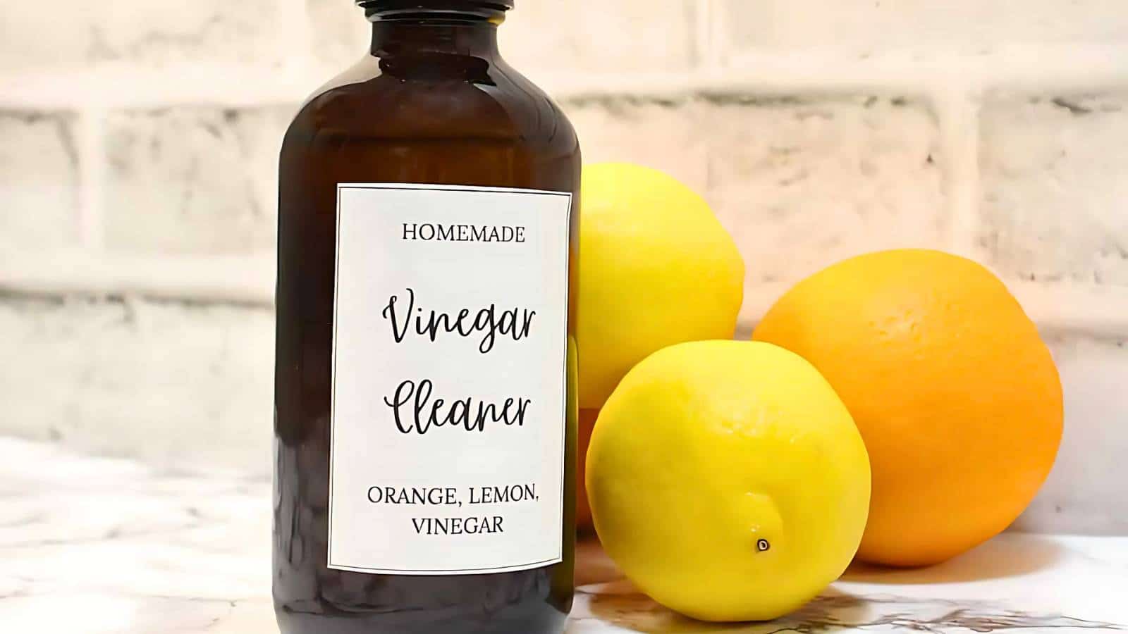 A bottle labeled "Homemade Vinegar Cleaner" sits on a marble surface, surrounded by a lemon, an orange, and another lemon, against a white brick background.