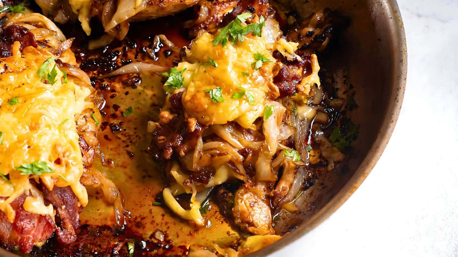 Close-up of a pan with cooked steak covered in melted cheese and garnished with chopped herbs, surrounded by caramelized onions.