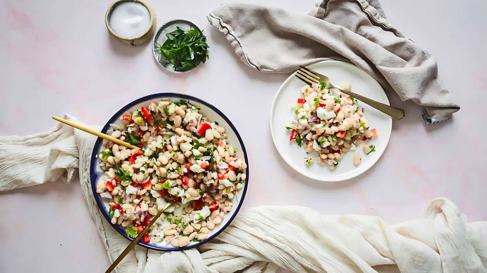 Two plates of bean salad with vegetables and herbs on a light tablecloth, garnished with parsley, alongside a small bowl of salt and a folded napkin.