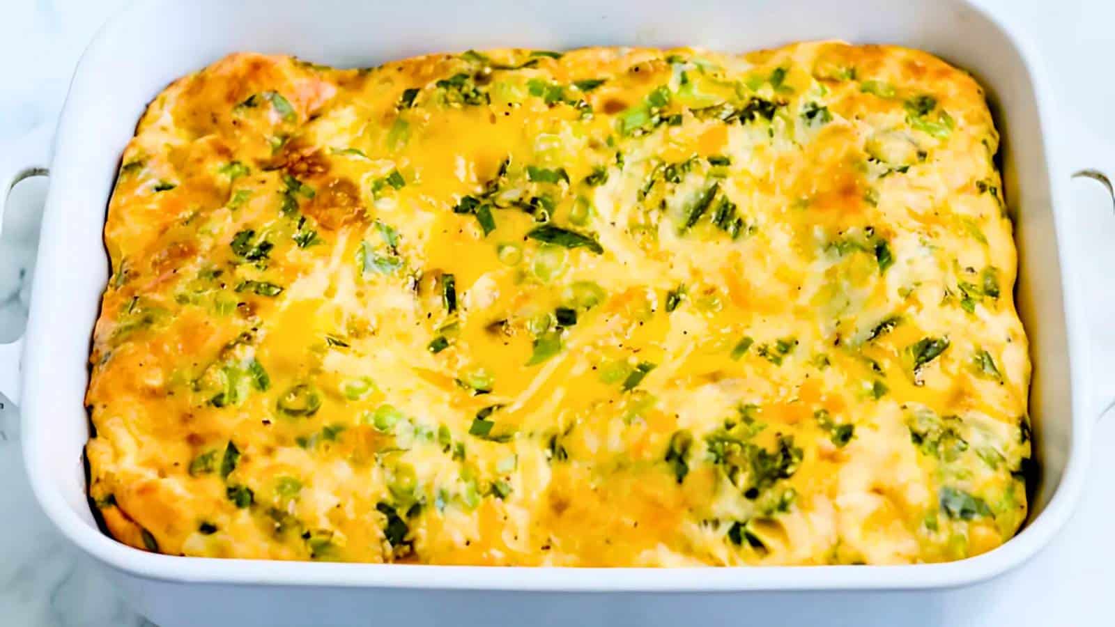 A baked egg casserole with melted cheese and green vegetables in a white rectangular dish.
