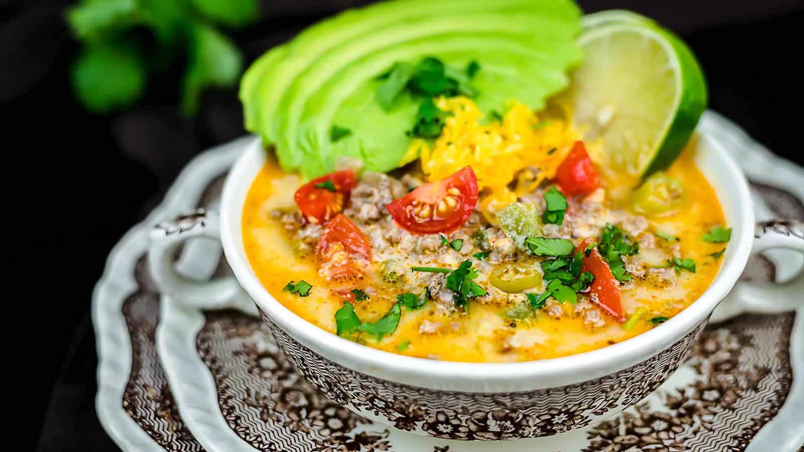 A bowl of soup topped with sliced avocado, shredded cheese, cherry tomatoes, cilantro, and a lime wedge.