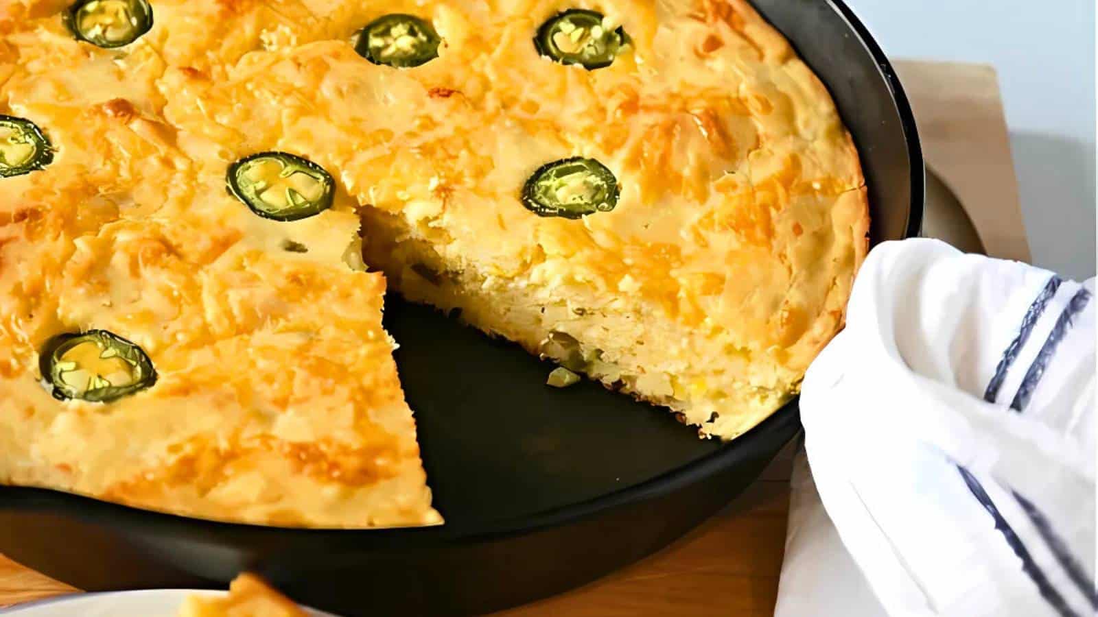 A slice of cornbread in a skillet with jalapeño slices on top. A white cloth napkin is placed nearby.