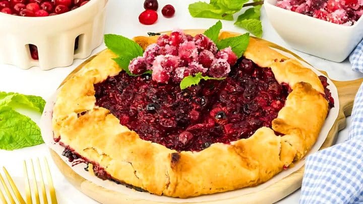 A rustic berry galette topped with sugared cranberries and mint leaves, surrounded by bowls of cranberries and a blue checkered cloth.
