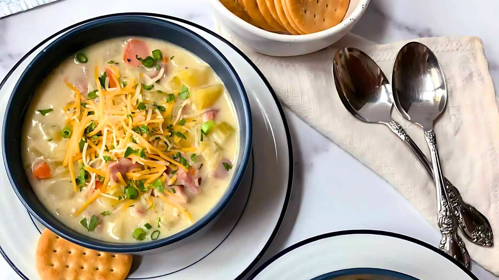 A bowl of creamy soup topped with shredded cheese and herbs, next to a plate of crackers and two spoons on a napkin.