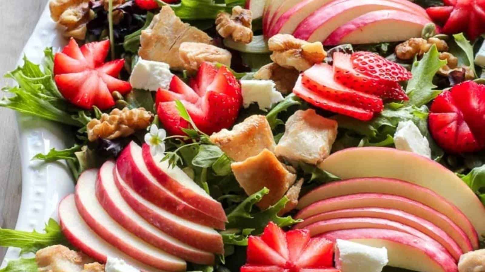 A salad with sliced apples, strawberries, chunks of chicken, walnuts, and feta cheese on a bed of mixed greens.