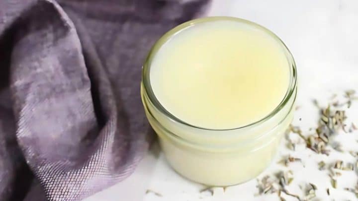 A glass jar filled with a creamy white substance is placed on a surface. A purple cloth is nearby along with scattered small dried flowers.