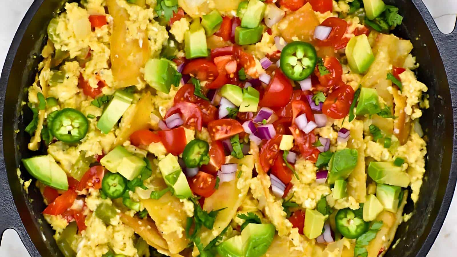 A skillet filled with scrambled eggs, avocado, tomatoes, jalapeño slices, red onion, and crispy tortilla pieces.
