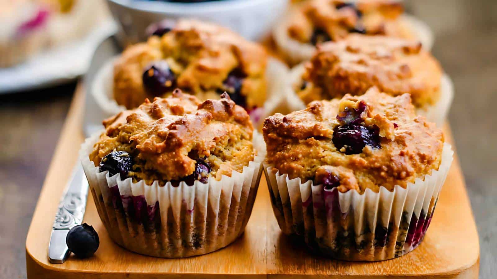 Four blueberry muffins on a wooden tray with a decorative knife beside them.