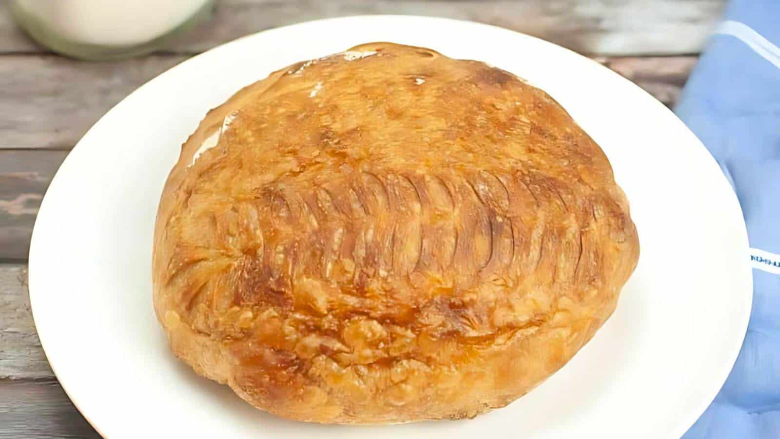 A round, golden-brown pie with a textured top is centered on a white plate. The background includes a blue cloth and a wooden surface.