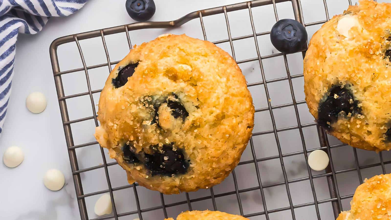 Blueberry muffins with sugar topping on a cooling rack, surrounded by scattered blueberries and white chocolate chips.