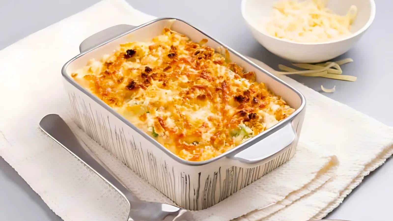 A baked dish in a rectangular casserole with a golden-brown crust, placed on a white cloth next to a bowl of shredded cheese and a serving spatula.