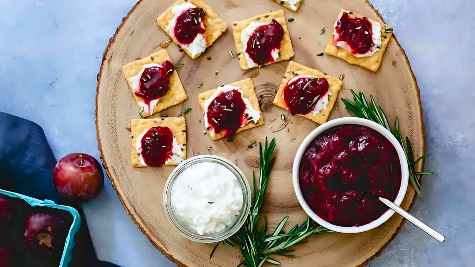 Wooden platter with crackers topped with cream and plum jam, a bowl of the jam with a spoon, a jar of cream, sprigs of rosemary, and whole plums beside a blue container.