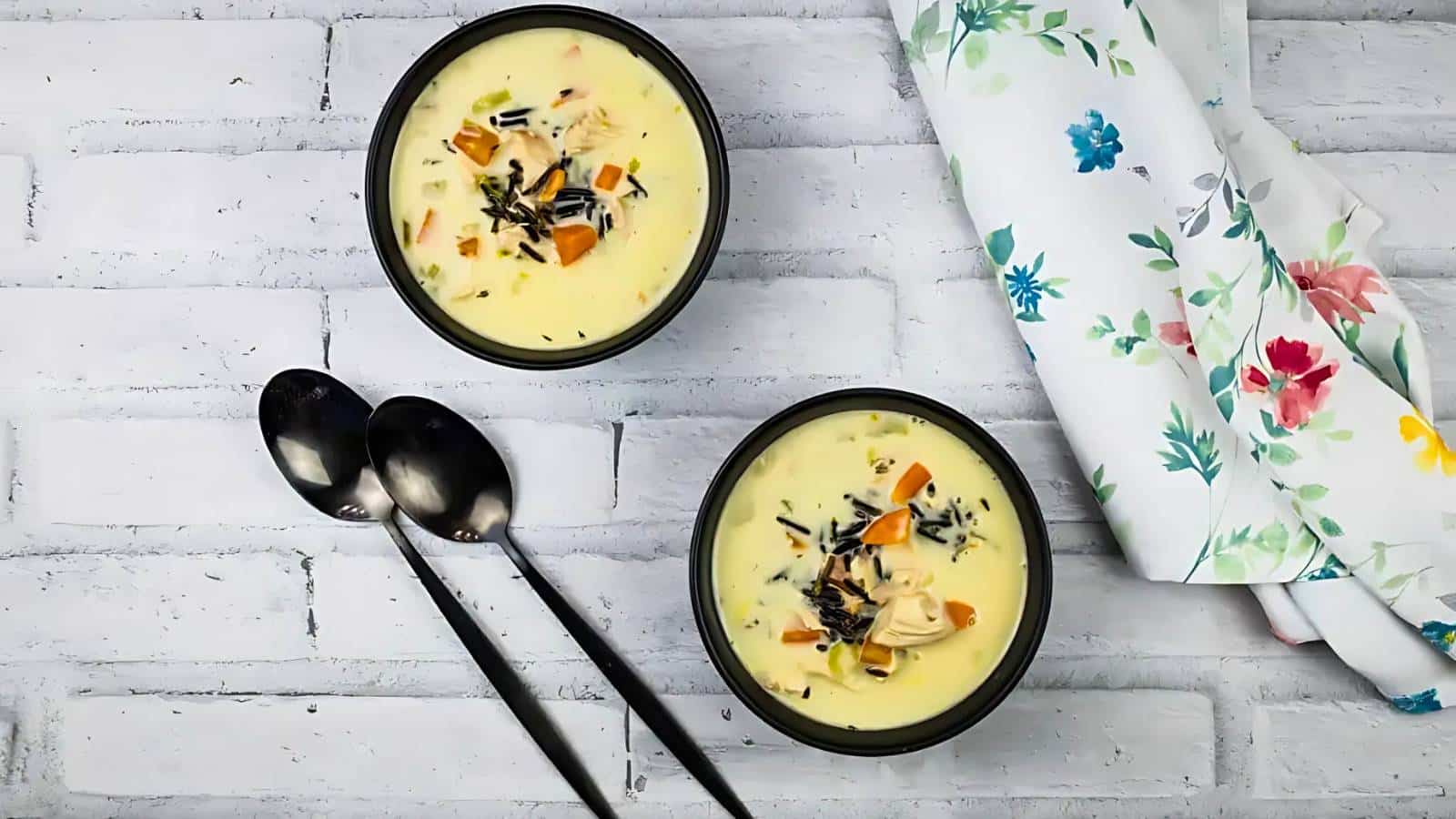 Two bowls of creamy soup with vegetables and herbs, placed on a white brick surface next to two spoons and a floral napkin.