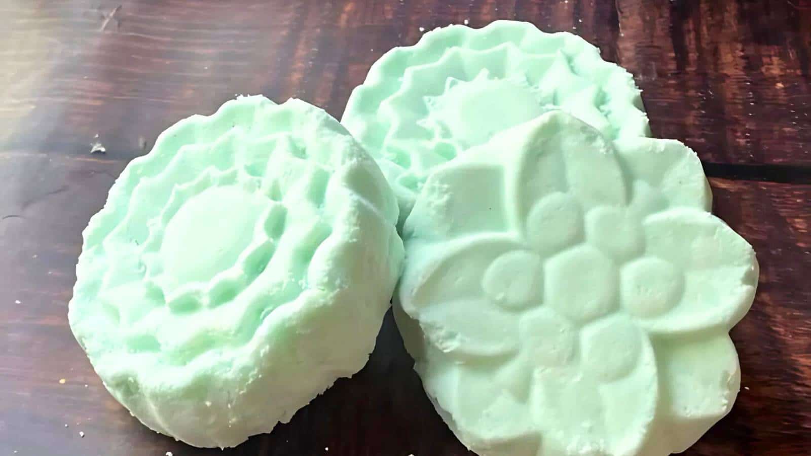 Three pastel green, intricately shaped soap bars on a wooden surface.