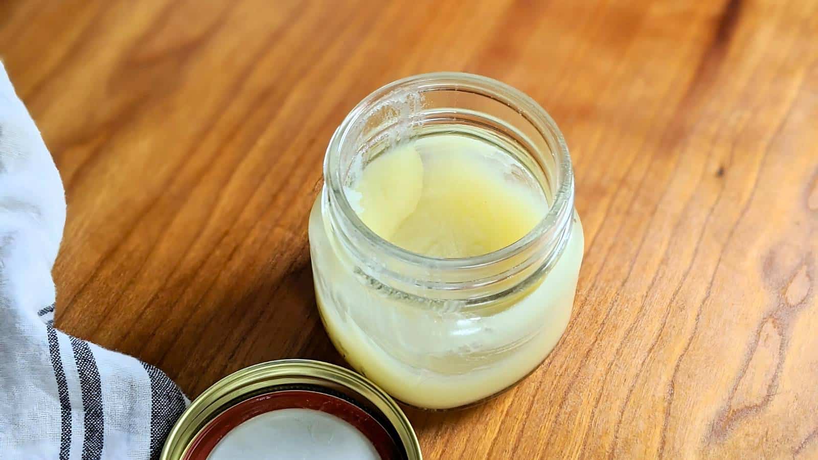 Open glass jar containing a light-colored cream on a wooden surface, with a lid and a cloth beside it.