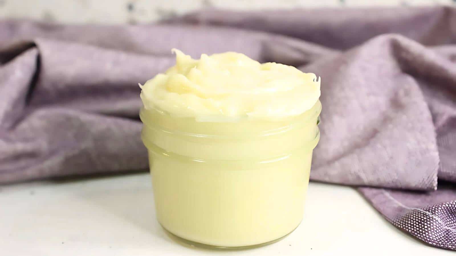 A small glass jar filled with creamy, pale yellow shea butter sits on a surface. A purple cloth is draped in the background.