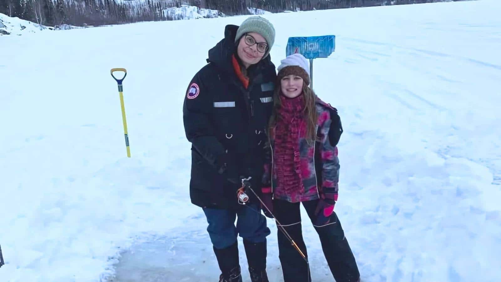 Two people smiling and standing in snow, dressed in winter clothing. A shovel and a sign are visible in the background.