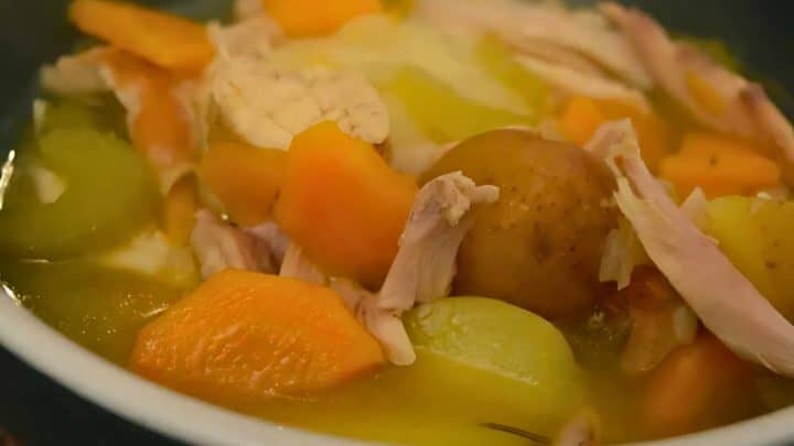 A bowl of soup containing chunks of chicken, carrots, potatoes, and other vegetables in a clear broth is the epitome of comfort foods.