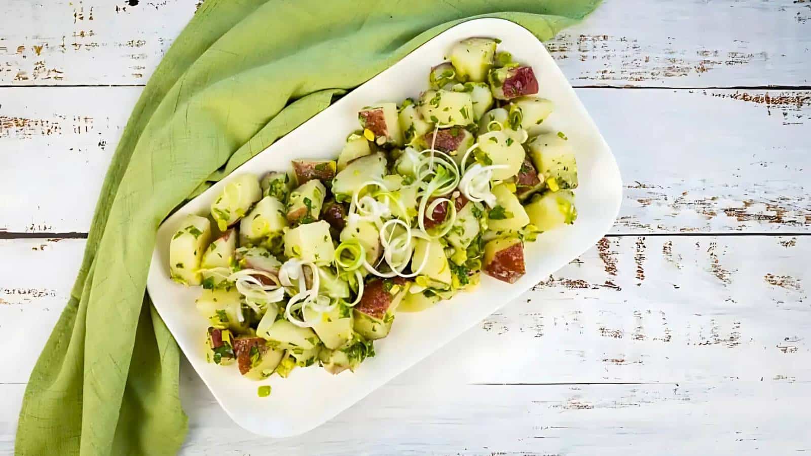 Plate of potato salad with chopped scallions and herbs on a wooden table, accompanied by a green cloth.