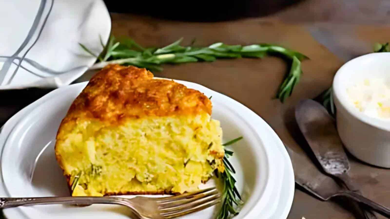 A slice of cornbread on a white plate with a fork, garnished with herbs. A white cloth and knife are nearby.