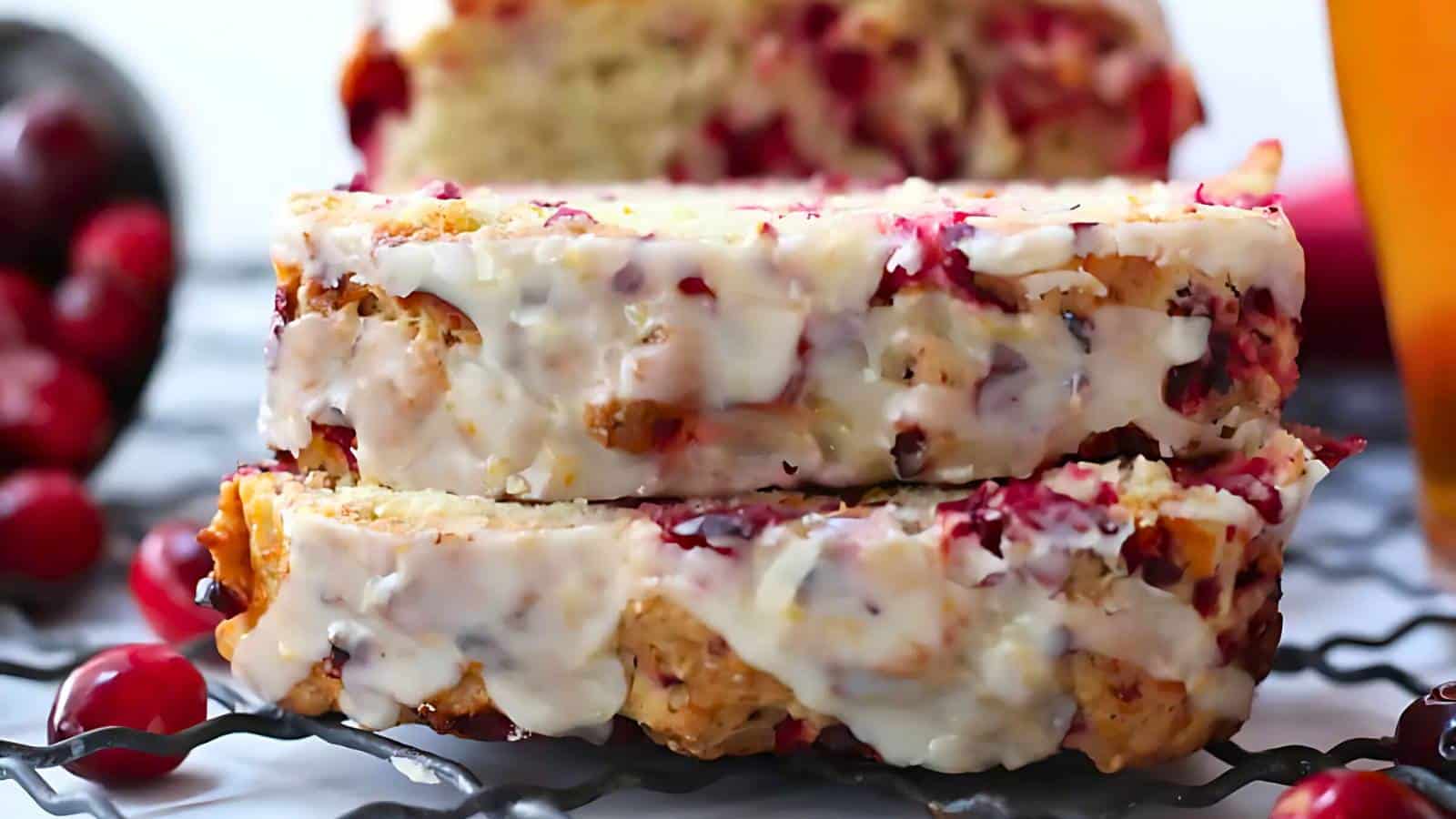 Two slices of cranberry bread with icing, placed on a cooling rack, surrounded by cranberries.