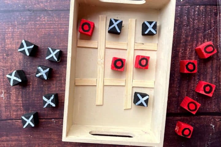 A wooden tic-tac-toe game with black and red blocks, displaying X's and O's. The game board is made of a small wooden tray with dividers. Some blocks are placed inside, while others are scattered outside.