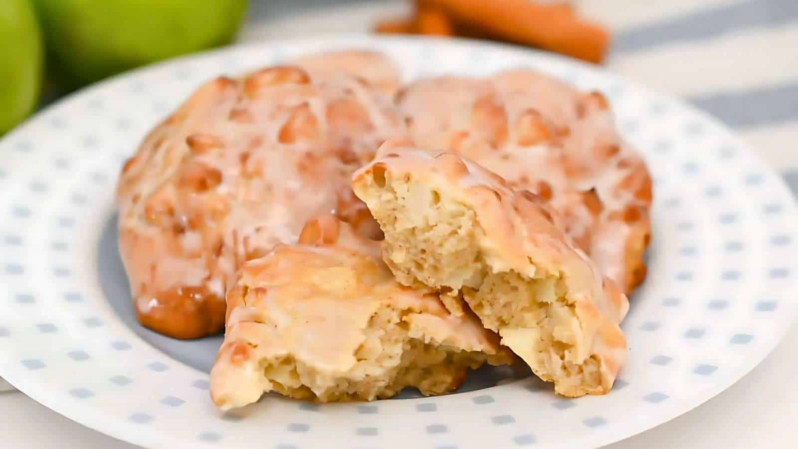 A white plate with apple fritters, one cut in half to show the inside. The fritters are glazed and sit on a dotted tablecloth.