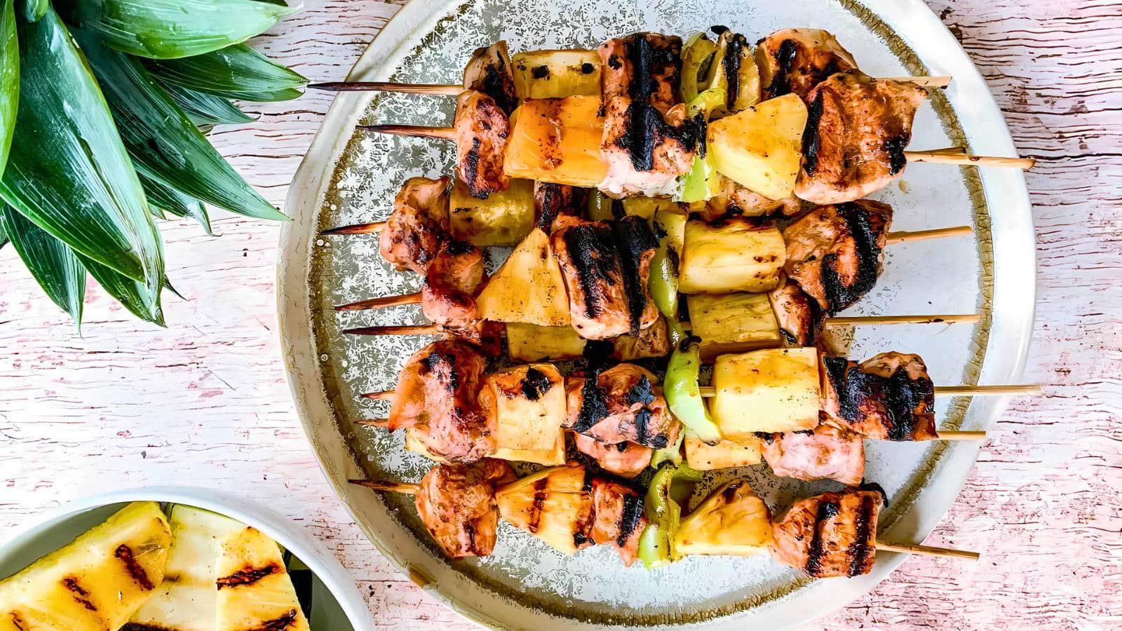 Grilled skewers with pineapple, meat, and peppers on a plate, with a side dish of pineapple and green leaves for decoration.