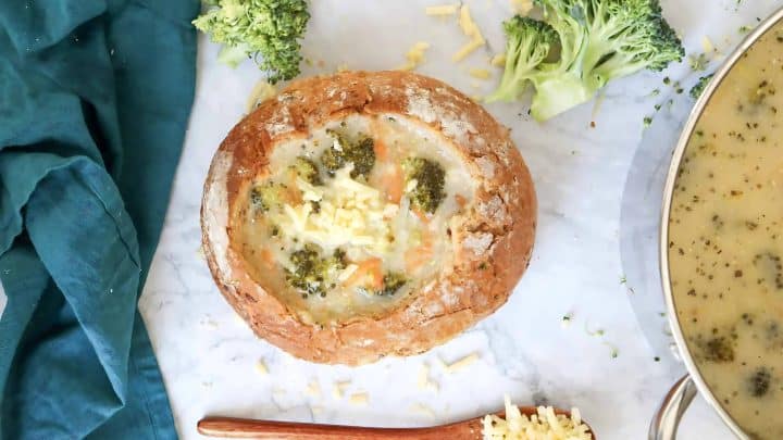 Broccoli cheese soup served in a bread bowl, surrounded by broccoli florets and shredded cheese on a marble surface.