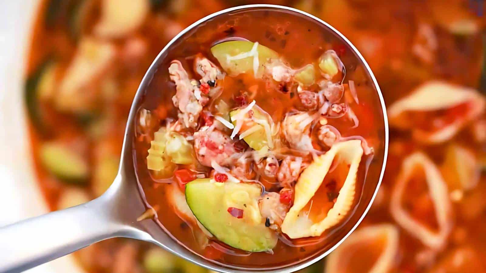 A ladle holding a serving of soup with pasta shells, ground meat, diced vegetables, and a tomato-based broth.