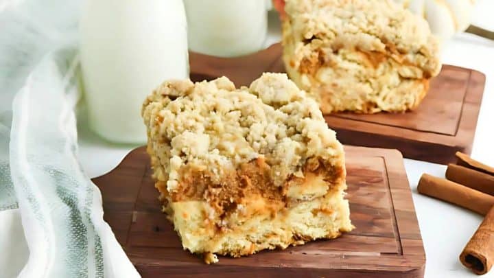 Two slices of crumbly apple coffee cake on wooden boards, with cinnamon sticks and milk bottles in the background.