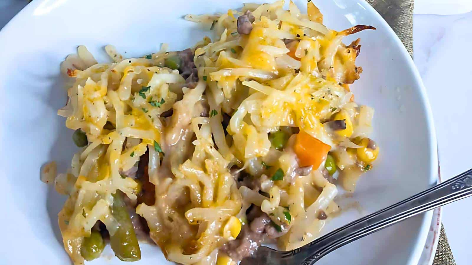 A white plate with a serving of hashbrown casserole mixed with cheese, vegetables, and ground meat. A fork rests on the plate's edge.