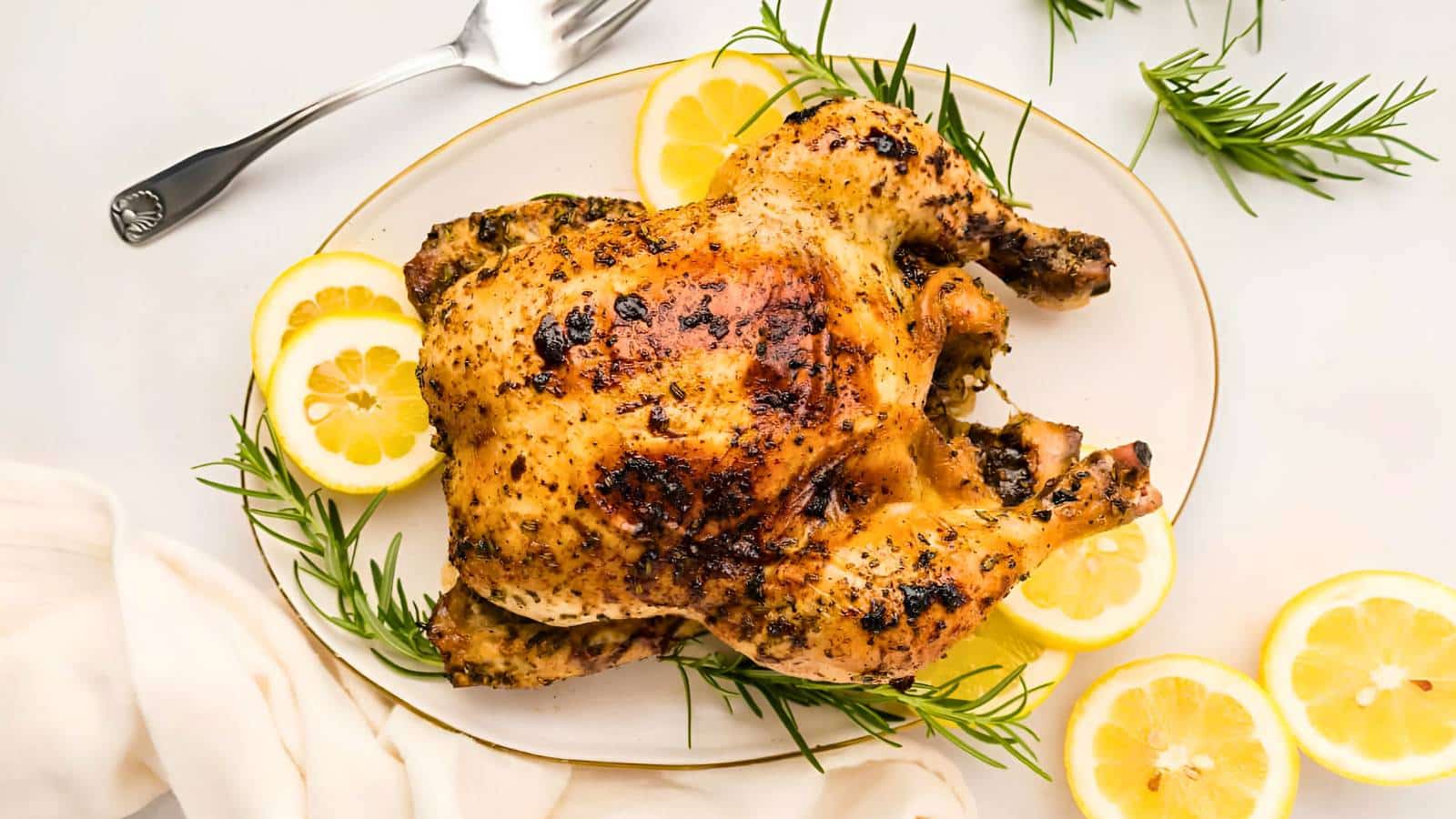 Roasted chicken on a platter garnished with lemon slices and rosemary, with a fork and napkin nearby.