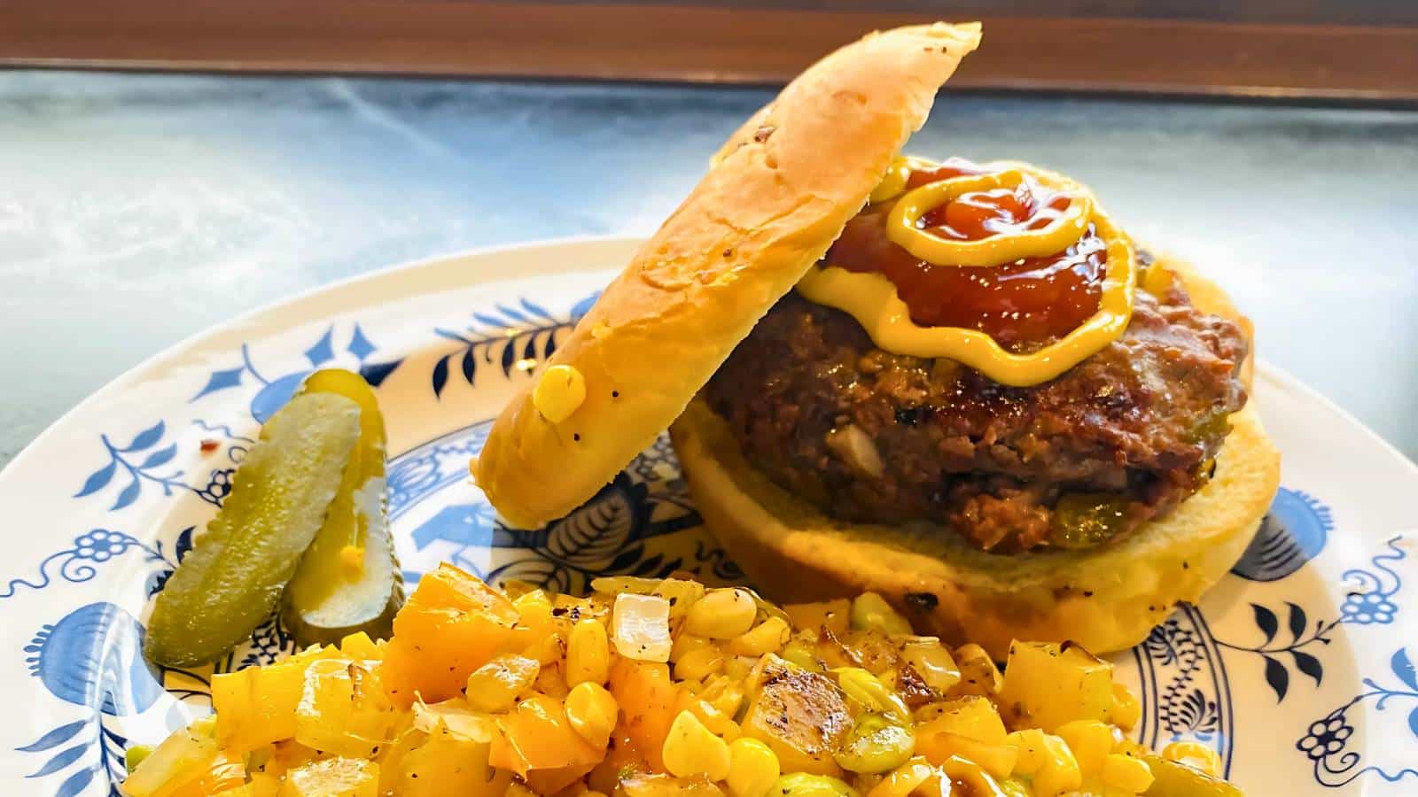 A hamburger with mustard and ketchup on a bun is served on a patterned plate with a side of diced potatoes, corn, and pickles.