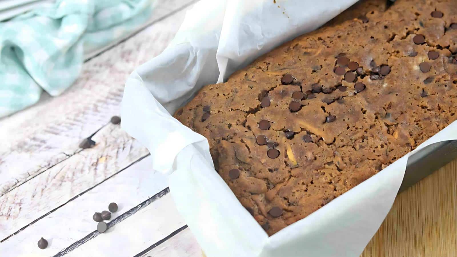 A loaf of chocolate chip bread sits in a parchment-lined baking dish on a wooden surface, with a few chocolate chips scattered nearby.