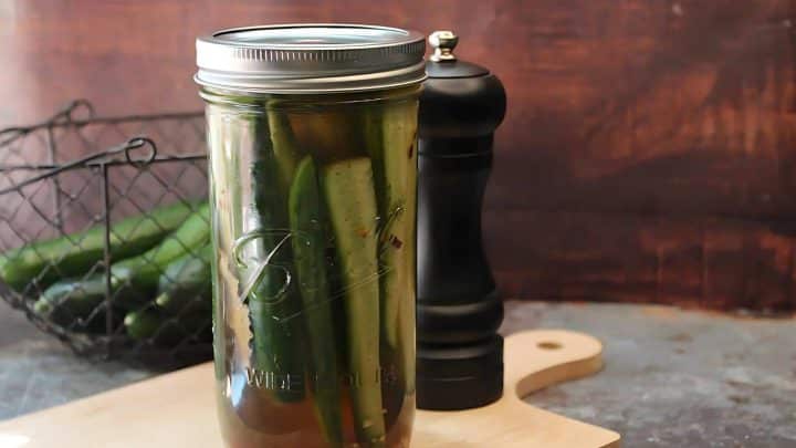 A jar of pickled cucumbers sits on a wooden board next to a black pepper grinder, with fresh cucumbers in a wire basket in the background.