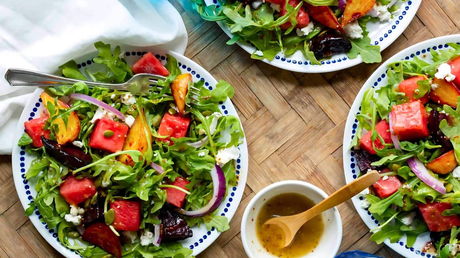 Three plates of fresh salad with watermelon, arugula, red onions, and other vegetables, surrounding a small bowl of dressing on a wooden table.