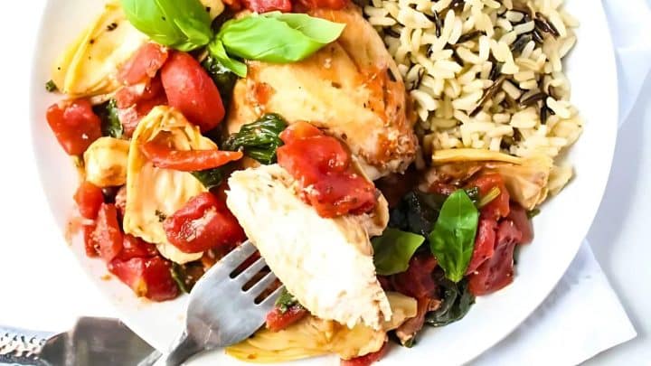 A plate with chicken breast, tomatoes, artichokes, fresh basil, and a side of wild rice. A fork rests on the plate.